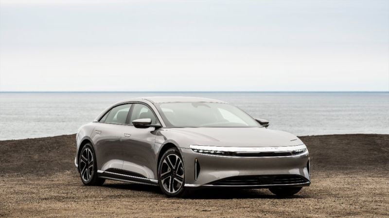 Silver 2025 Lucid Air electric sedan parked on rocky terrain with calm ocean horizon in background, sleek aerodynamic profile and distinctive LED headlights
