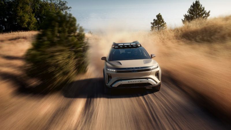 Silver Lucid Gravity electric SUV shown in dynamic three-quarter front view, kicking up dust while driving on dirt road, featuring distinctive LED lighting and roof rack.
