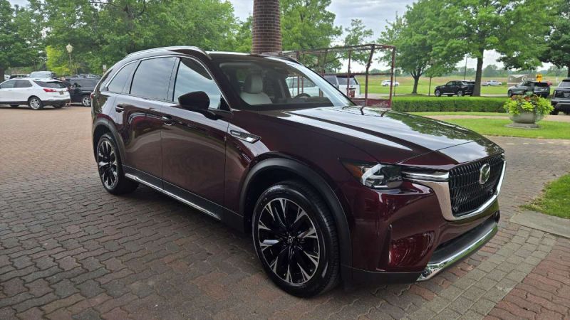 Deep burgundy 2025 Mazda CX90 SUV parked on brick pavement, angled front three-quarter view with lush green trees in background.