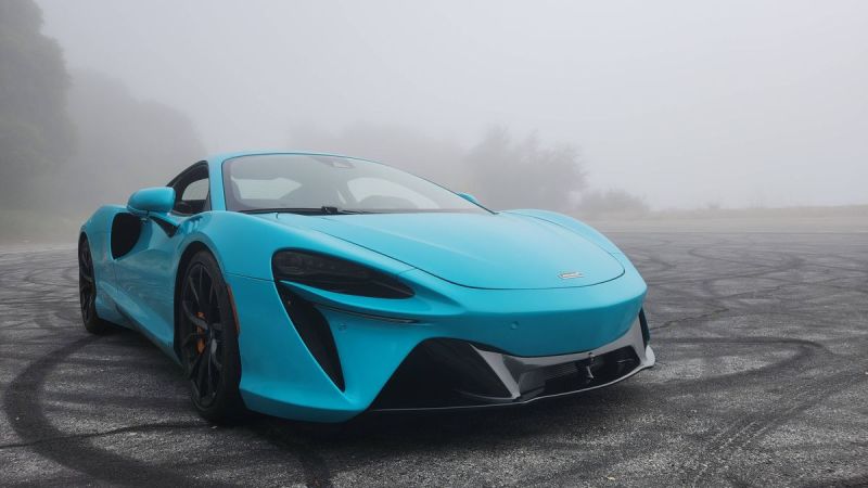 A turquoise 2025 McLaren Artura parked on a misty road, showcasing sleek lines and bold black wheels.
