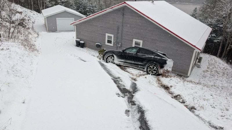 Black Ford Mustang Mach-E parked on a snowy driveway beside a house, showing winter driving conditions and snow-covered landscape.