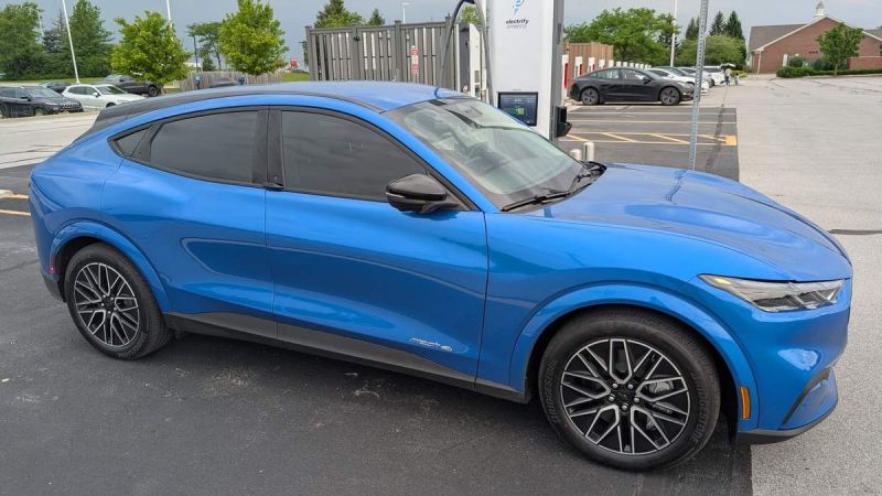 A blue Ford Mustang Mach-E parked near an electric vehicle charging station, surrounded by trees and other cars.