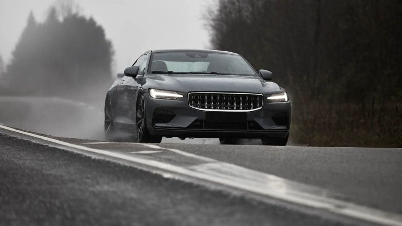 A sleek gray car drives through a misty landscape, splashing water on a wet road, showcasing its dynamic design and performance.