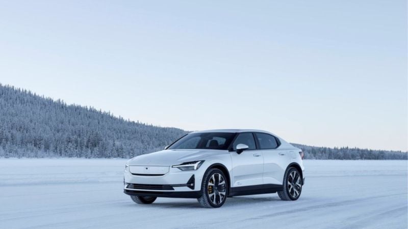 A sleek white electric car parked on a snow-covered landscape, surrounded by snow-laden trees under a clear sky.