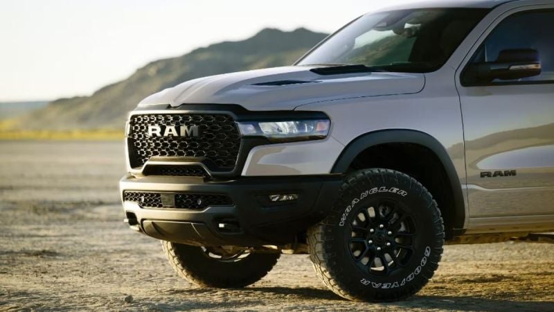 A silver 2025 RAM 1500 pickup truck shown from the front quarter angle, featuring black off-road wheels, aggressive honeycomb grille design, and rugged front bumper against a mountainous backdrop.