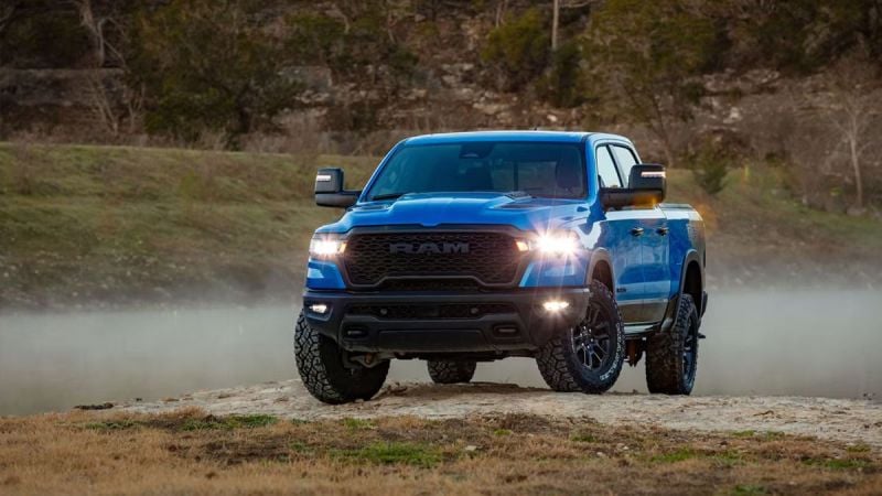 A bright blue 2025 Ram 1500 pickup truck shown from a front three-quarter angle, with LED headlights illuminated and off-road tires kicking up dust on rugged terrain.