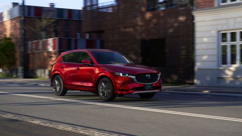 A 2025 red Mazda CX-5 cruising down a city street, showcasing modern architecture and vibrant colors in the background.