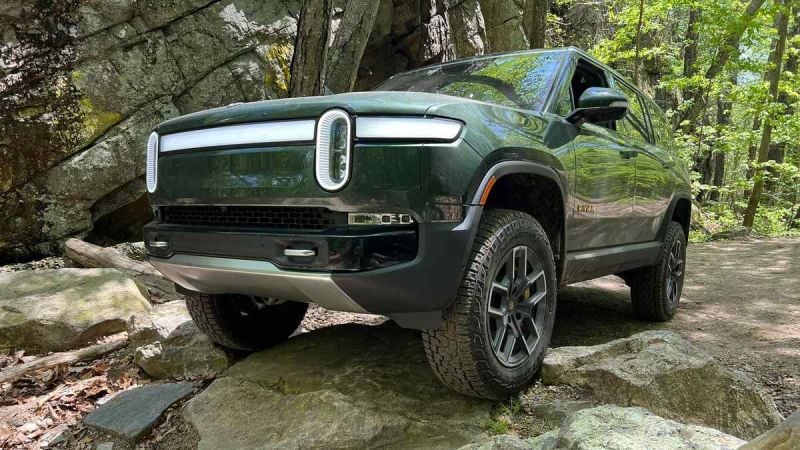 A dark green Rivian R1S electric SUV shown from the front quarter angle, demonstrating its off-road capabilities while navigating rocky terrain in a wooded setting.