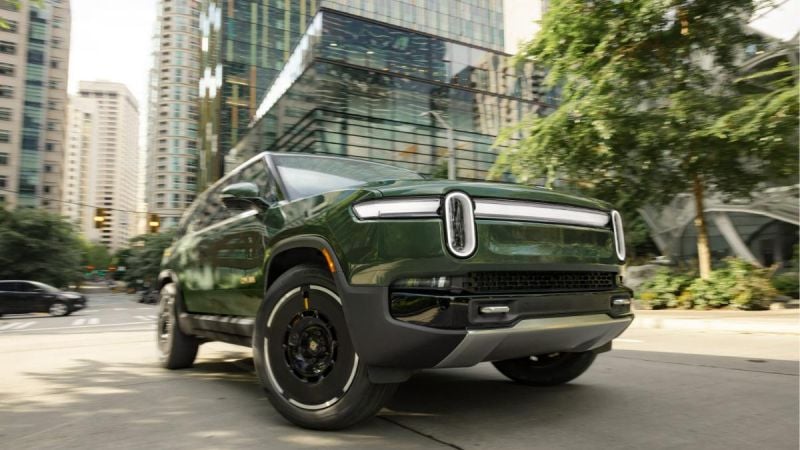 Low-angle view of a green 2025 Rivian R1S electric SUV driving through a modern city street with glass buildings