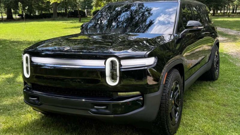 A sleek black electric SUV with distinctive round headlights and a glossy finish, parked on a grassy area surrounded by trees.