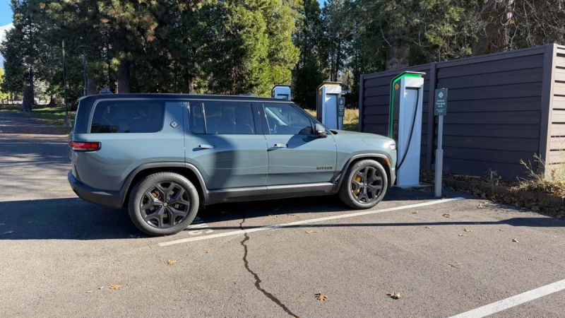 A silver 2025 Rivian R1S is shown from the rear three-quarter view at an EV charging station, featuring black accent wheels and trim.