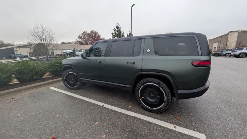 Forest Green 2025 Rivian R1S SUV photographed from a rear three-quarter angle in a parking lot, showing its boxy design, black wheels, and matte finish.
