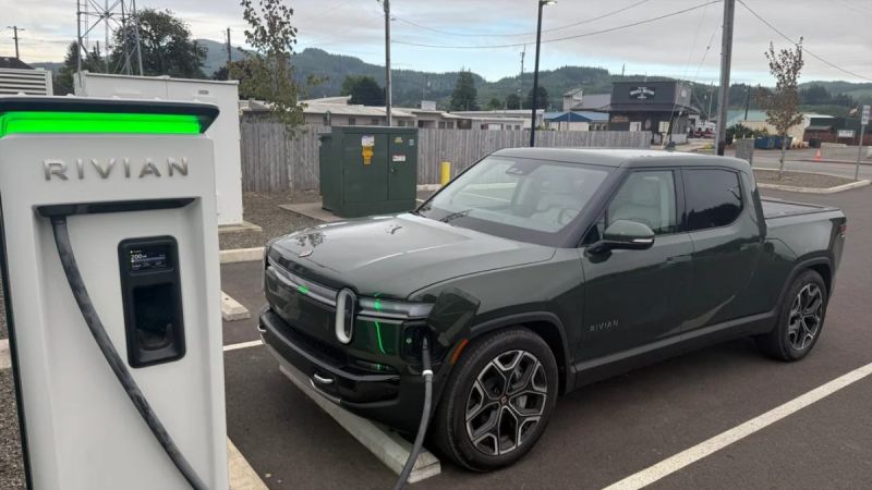 Dark green Rivian R1T electric pickup truck charging at a white Rivian charging station in an overcast suburban parking lot with mountains in background