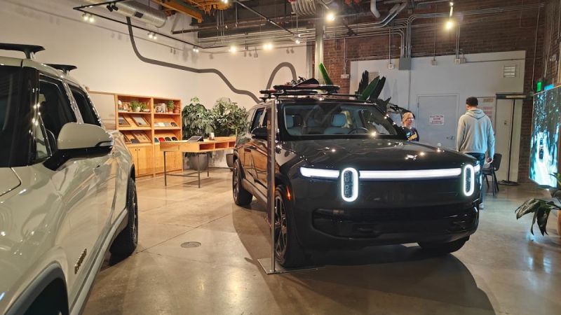 2025 Rivian R1S electric SUV in black, front three-quarter view, displaying distinctive LED headlight bar, shown in industrial-style showroom with exposed brick walls.