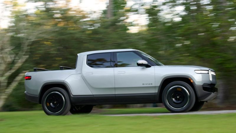 Photograph of the 2025 Rivian R1T electric truck in a gray color, captured from a three-quarter front angle with trees visible in the background.