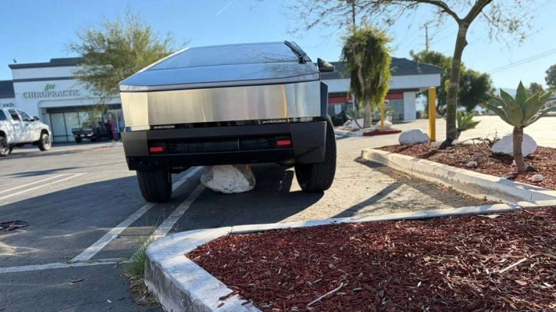 Tesla Cybertruck parked in a commercial parking lot with a large white boulder lodged under its front end, showing the electric truck elevated slightly off the ground.