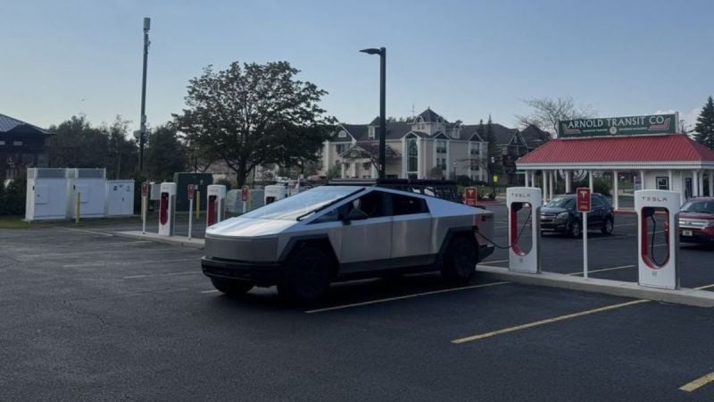 A Tesla Cybertruck charging at a station, surrounded by other chargers and vehicles, set against a backdrop of trees and buildings.