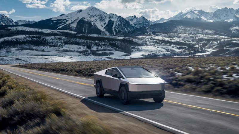 A futuristic electric truck drives along a scenic road with snow-capped mountains and a clear blue sky in the background.