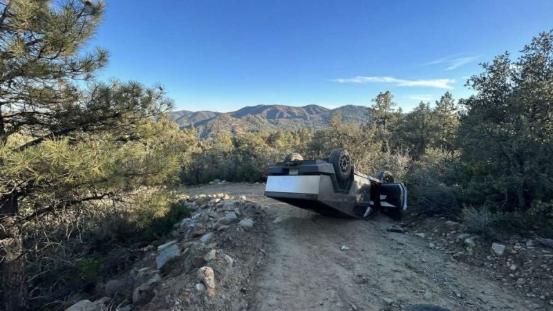 Tesla Cybertruck overturned on a narrow mountain dirt road surrounded by pine trees and rocky terrain
