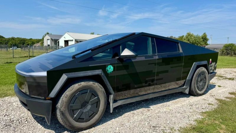 Black futuristic truck parked on gravel near a fenced grassy area and buildings.