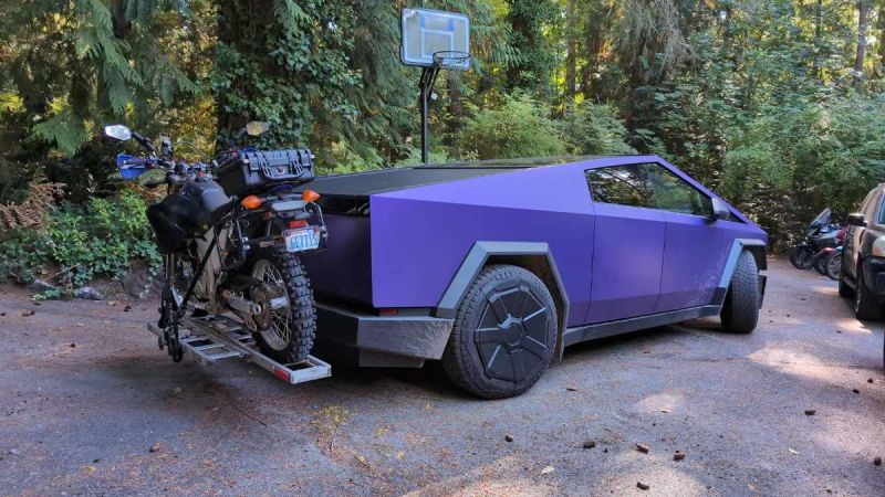 Purple futuristic truck with a motorcycle mounted on the back, parked in a wooded area.