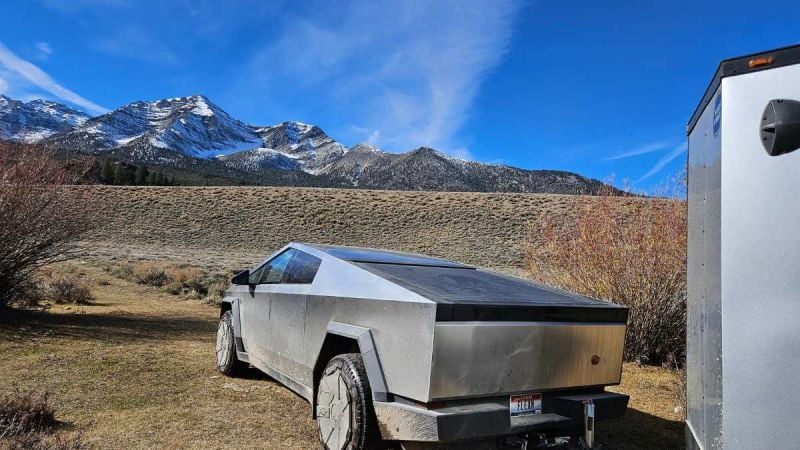 A Tesla Cybertruck in silver/stainless steel finish is parked in a mountainous landscape, shown from a rear three-quarter angle with snow-capped peaks visible in the background.