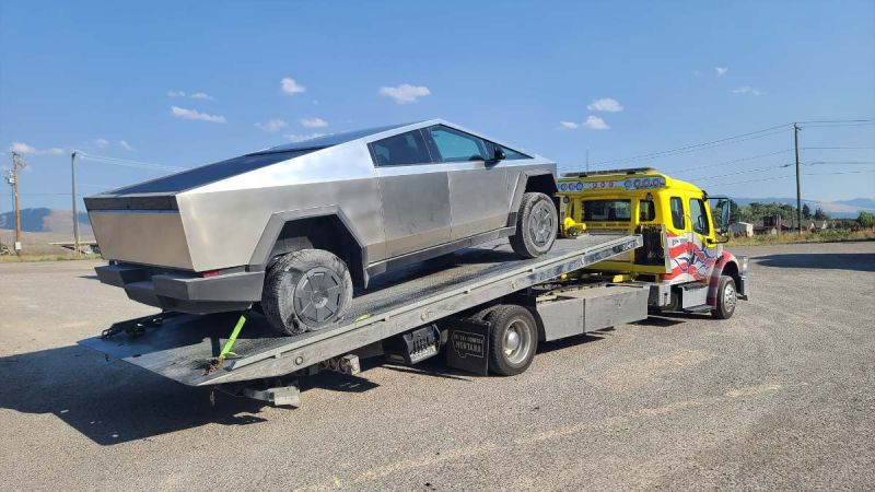 Stainless steel 2025 Tesla Cybertruck loaded on bright yellow tow truck against rural mountain backdrop