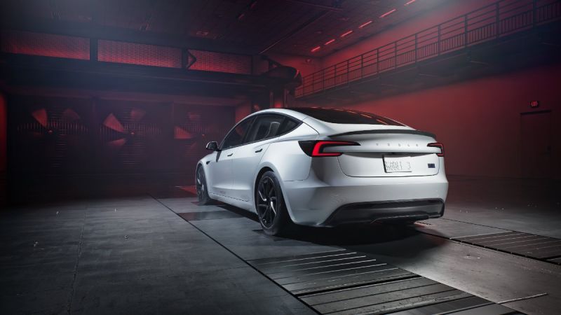 Rear three-quarter view of a white Tesla Model 3 Highland in an industrial wind tunnel testing environment with dramatic red lighting. The sleek aerodynamic design is highlighted by performance tires and minimalist rear end.