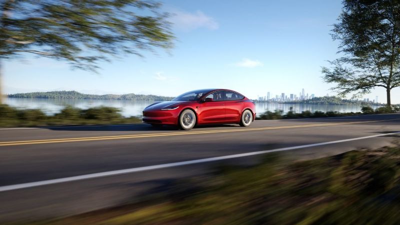 Red car driving on a road beside a lake, with a city skyline in the background.
