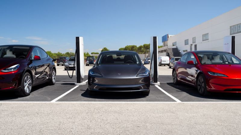 Multiple Tesla Model 3 vehicles in gray and red parked at charging stations, with white Supercharger pedestals visible, outside a modern commercial building.