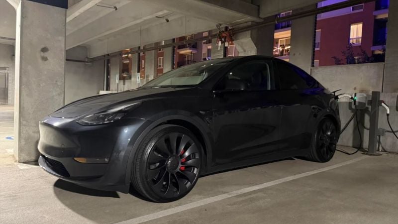 Midnight black 2025 Tesla Model Y charging in concrete parking garage, aggressive profile with black turbine wheels at night