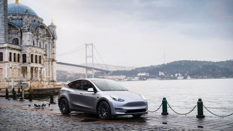 A silver Tesla Model Y parked by a historic building along a waterfront, with a bridge and misty hills in the background.