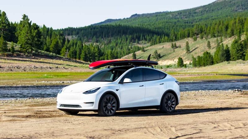 A white Tesla Model Y parked on sandy terrain by a river, with a red surfboard secured on its roof against a backdrop of green hills.