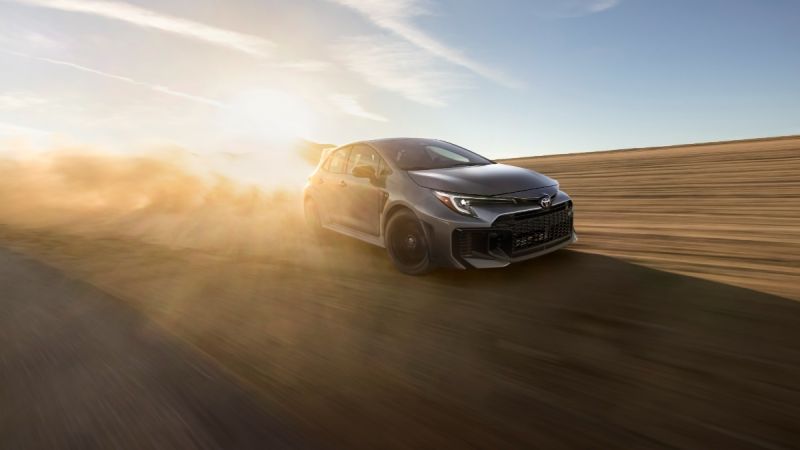 2023 Toyota Corolla in metallic gray, shown in three-quarter front view, driving on desert road at sunset with dramatic lighting and dust effects.