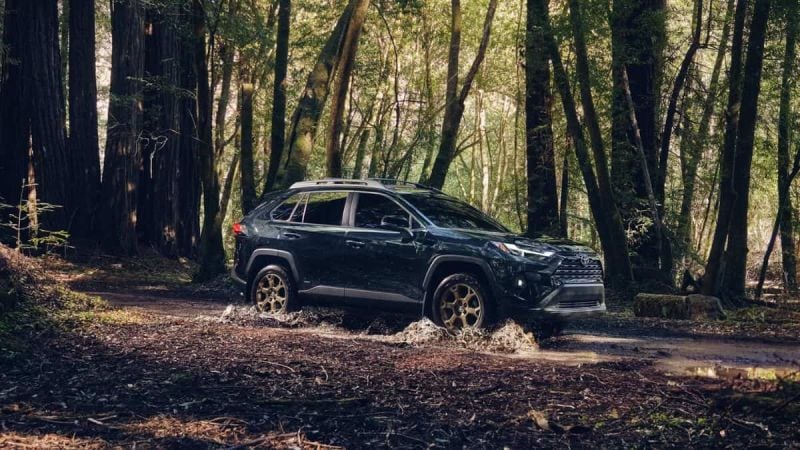 A dark Toyota RAV4 drives through a mud puddle on a forested dirt path surrounded by tall and sunlit trees, conveying adventure and ruggedness.