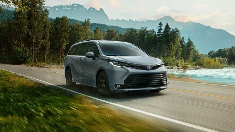 Silver 2025 Toyota Sienna minivan shown from a front three-quarter view, driving on a scenic mountain road with snow-capped peaks and turquoise water in the background.