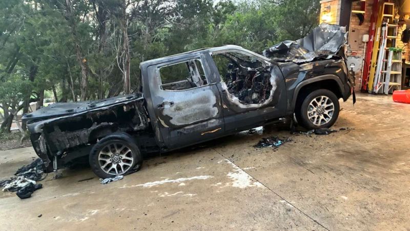 Severely fire-damaged black Toyota Tundra with melted rear section, parked outside brick building. Chrome wheels remain intact despite extensive burn damage.
