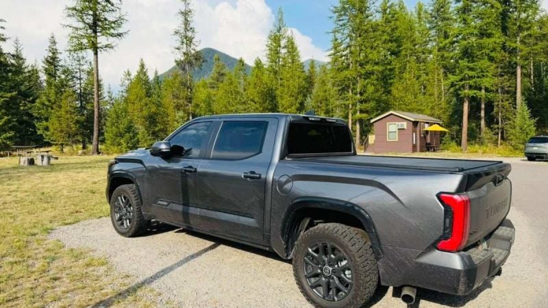 A dark gray/charcoal 2025 Toyota Tundra pickup truck shown from the rear quarter view, parked in a scenic mountain setting with evergreen trees and cabins in the background.