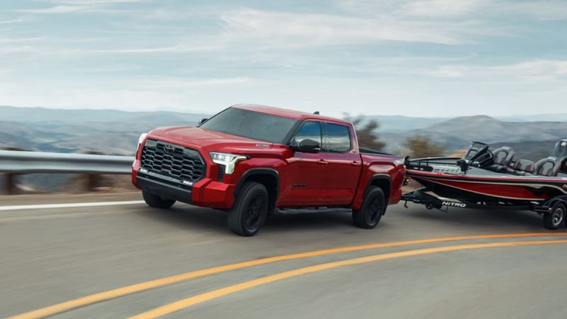 Red Toyota Tundra towing a boat on a winding mountain road. The backdrop shows a scenic landscape of rolling hills under a partly cloudy sky.