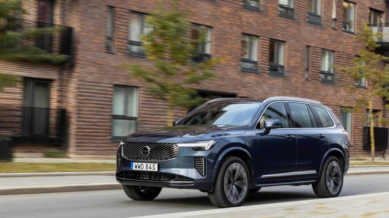A 2025 blue Volvo CX90, speeding past modern brick buildings, showcasing its stylish design and dynamic performance.