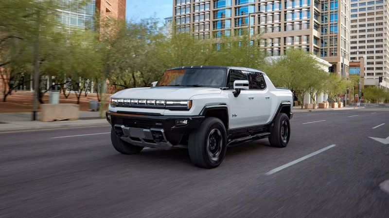 A white 2025 GMC Hummer EV drives through a city street with modern buildings and greenery in the background, showcasing its rugged design.