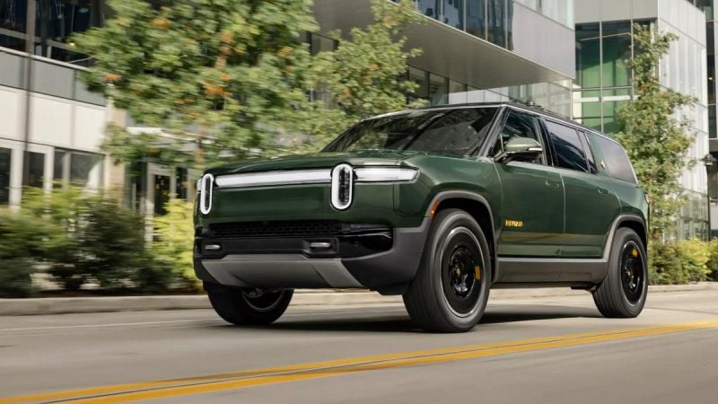 Green Rivian R1S electric SUV driving through a modern city street with glass office buildings in the background