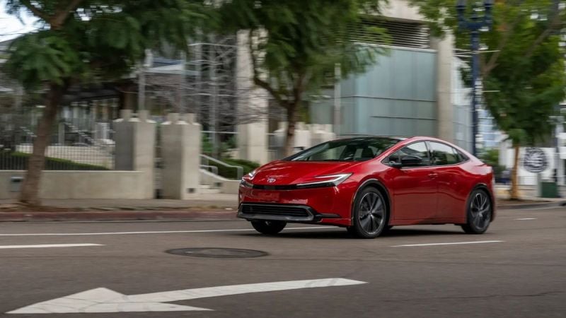 Red 2025 Toyota Prius driving through an urban street, showcasing sporty exterior styling and updated front fascia.