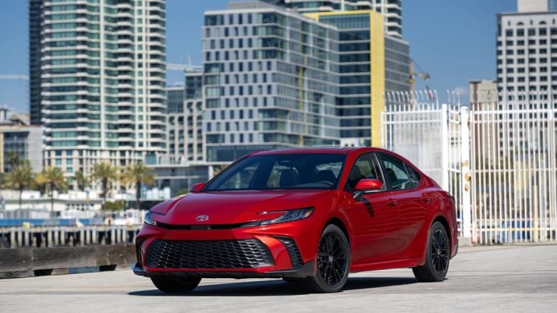A sleek red Toyota sedan parked in an urban setting with modern skyscrapers and palm trees in the background.