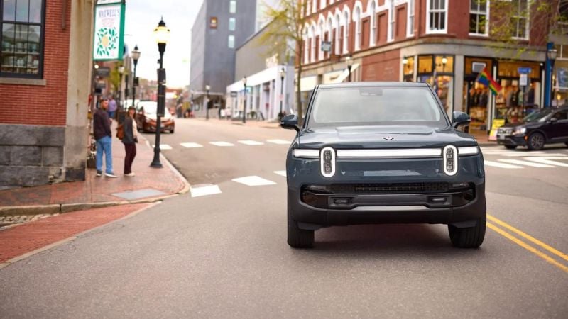 Front view of a Rivian R1S electric SUV driving through an urban downtown street, featuring signature vertical LED headlights and a boxy adventure-focused design.