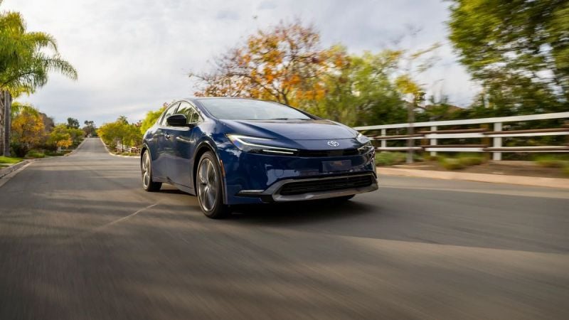 2025 Toyota Prius Limited in Reservoir Blue driving down a suburban street, front three-quarter view with motion blur and trees lining the road.