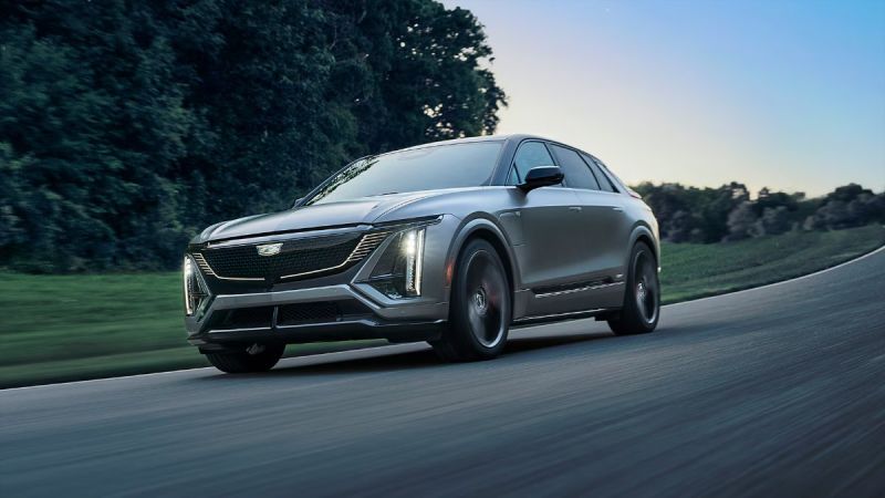 Silver 2026 Cadillac Lyriq electric SUV curving on a road with green trees and blurred motion