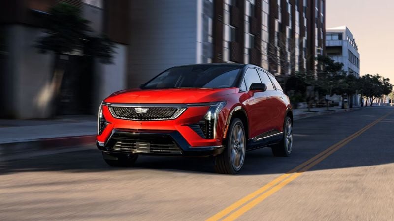 Red SUV driving down a city street with buildings and trees in the background.