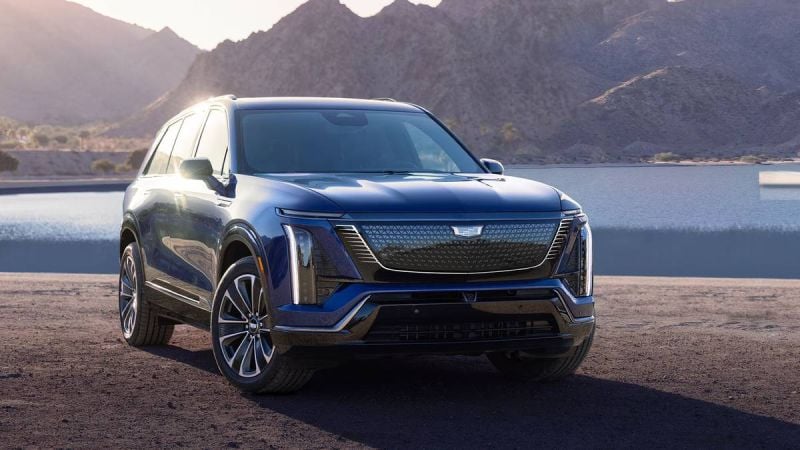 A sleek, dark blue 2026 Cadillac Vistiq parked by a serene lake, with mountains in the background and a sailboat on the water.
