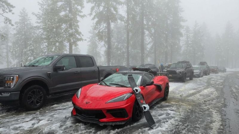 Red Chevrolet Corvette C8 parked in a snowy mountain lot with a pair of skis leaning on the front bumper, surrounded by snow-covered trucks and trees.